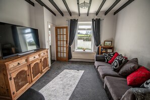 Un salon avec un téléviseur posé sur un meuble en bois, un canapé gris agrémenté de coussins à motifs et une vue sur un jardin, visible à travers une fenêtre ornée de rideaux.