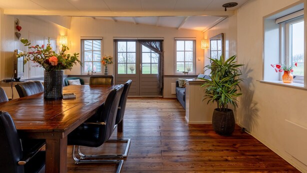 Ein Essbereich mit Holztisch, schwarzen St ühlen und einer Blumenvase. Es gibt Fenster mit Vorhängen, eine Pflanze und einen Blick ins Freie.