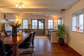 Un coin repas avec une table en bois, des chaises noires et un vase de fleurs. On y trouve des fenêtres avec des rideaux, une plante et une vue sur l’extérieur.