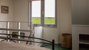 Ein Zimmer mit einem Bett, einem Fenster mit Blick auf ein grünes Feld, einer Holztür und einem Korb auf dem Boden.