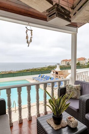 A covered outdoor area with a pool, lounge chairs, and a view of the sea.