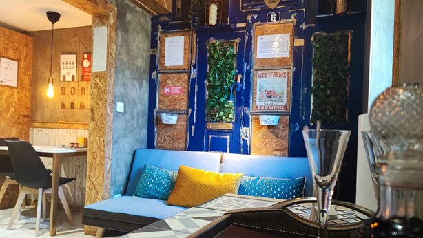 A cozy seating area with a blue sofa and cushions, a wooden table with a glass, and a wall decorated with various signs and plants.