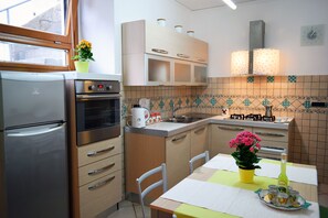 A compact kitchen with a refrigerator, oven, microwave, and a sink. There are cabinets, a table with chairs, and a potted plant on the windowsill.