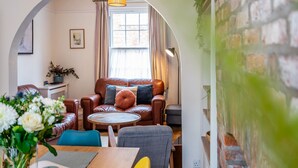 A cozy living room with a brown leather sofa, a round coffee table, and a blue chair. There is a window with curtains, a lamp, and a plant on the wall. - Buckingham Townhouse -  a family break that sleeps 6 guests in 3 bedrooms (York)