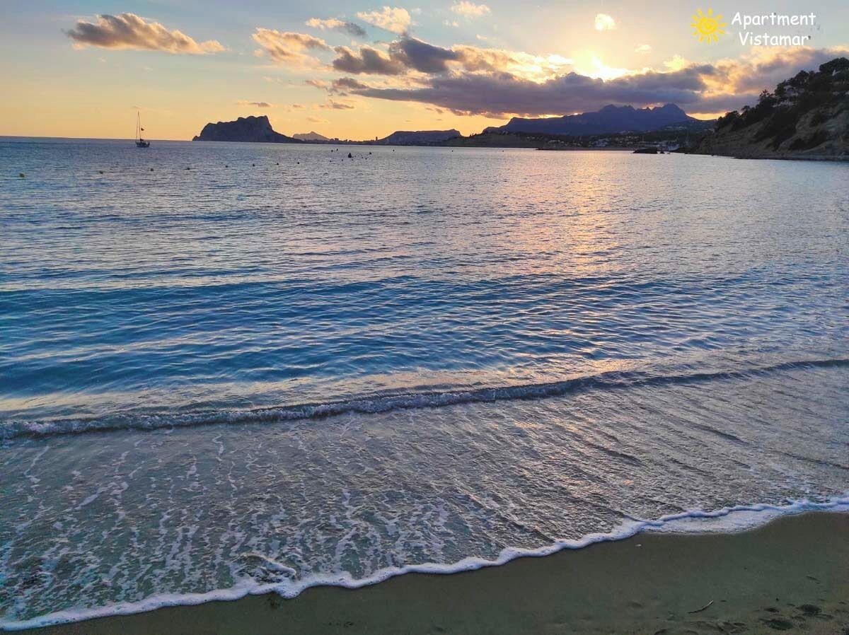 A beach with gentle waves, a sailboat, and distant mountains under a sunset sky.
