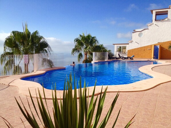 A swimming pool with a clear view of the ocean, surrounded by palm trees and a white building with a balcony.
