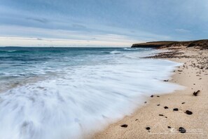 A beach with waves gently lapping at the shore, a rocky outcrop in the distance, and a clear sky.