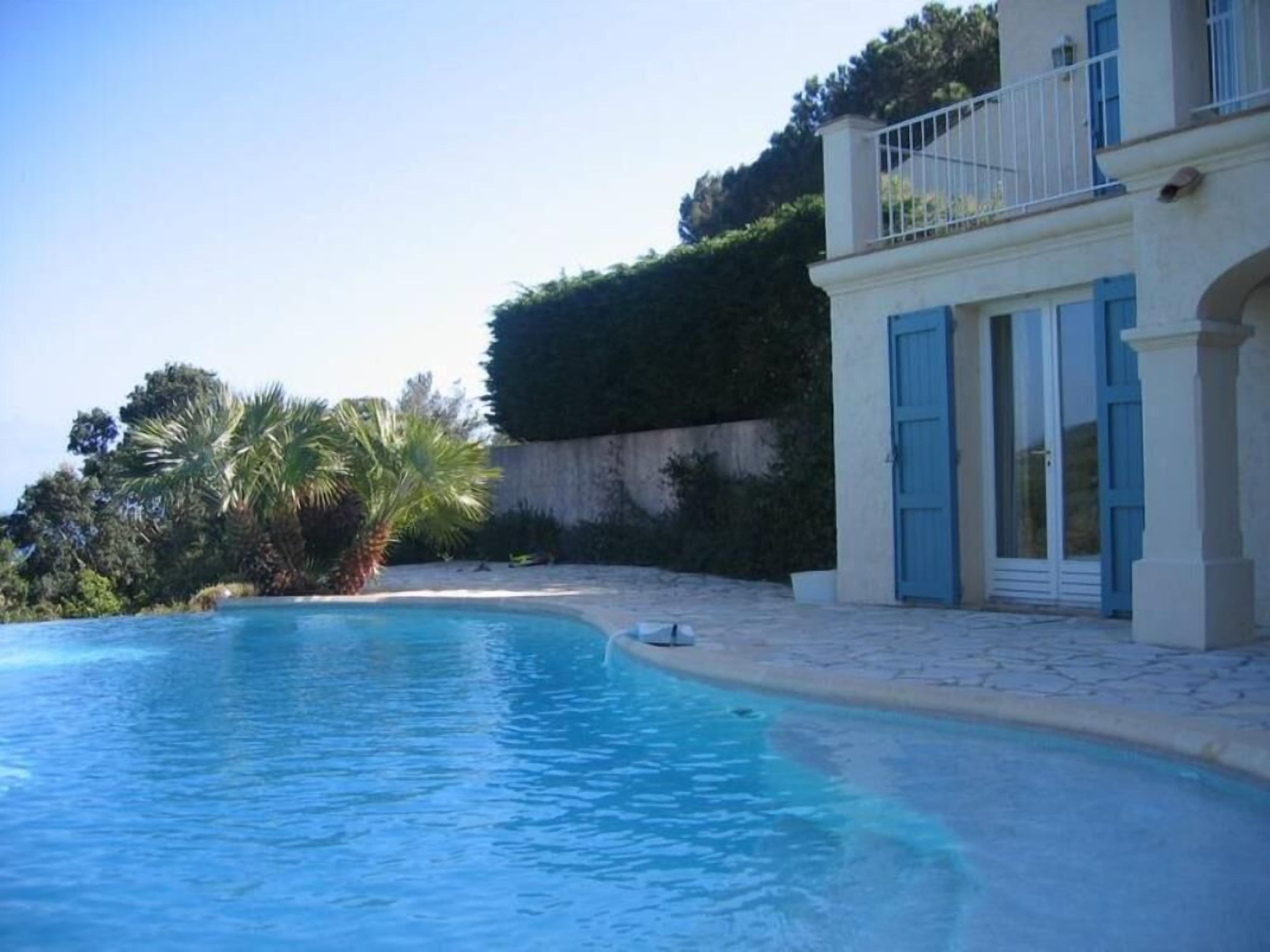 A swimming pool with a curved edge, a building with blue shutters and a balcony, and a hedge in the background.
