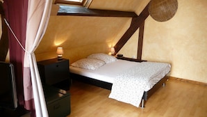 Un dormitorio con techo inclinado, vigas de madera, una cama con colcha blanca, dos lámparas de noche y una maleta en el suelo.