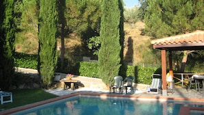 Une piscine agrémentée d’une pergola et de chaises, entourée de grands arbres et d’un mur de pierre.
