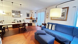 Ein Wohnzimmer mit blauem Sofa, einer Essecke mit Tisch und Stühlen sowie einer Küchenzeile mit Herd und Fenster.