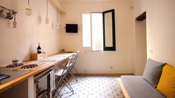 A compact kitchen with a wooden countertop, a dining table, and chairs. A television is mounted on the wall, and a window with a black frame is visible.