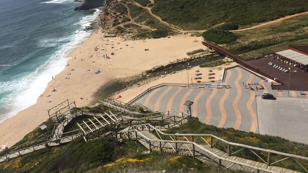 Ein Küstenstrand mit einer Holztreppe, die zum Ufer führt, feinem Sand und einer Pflasterfläche mit Sitzgelegenheiten.