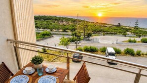 Ein Balkon mit Tisch für zwei Personen, mit Blick auf einen Parkplatz und den Sonnenuntergang über dem Meer.