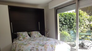 Ein Schlafzimmer mit einem Bett, einem dunklen Kopfteil und Blick auf eine Terrasse mit Stühlen und Pflanzen.