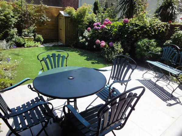 Une terrasse avec une table ronde et quatre chaises, entourée d’un jardin où poussent différentes plantes et fleurs.