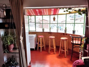 Cozy kitchen with a large window offering a view of the residential street, featuring a wooden countertop, refrigerator, and stools with cushions.