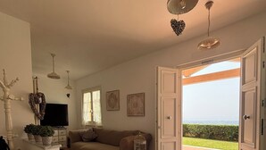 Un vaste salon doté d’un plancher en bois, d’un canapé beige, d’une table basse blanche et d’une cage à oiseaux. Des luminaires sont fixés au plafond, et une porte donne accès à un balcon avec vue.