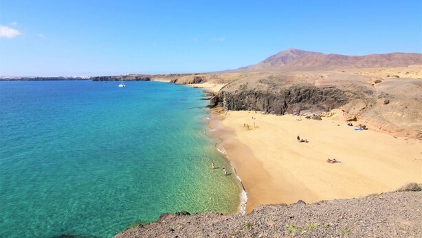 A coastal beach with clear turquoise water, sandy shore, and rocky cliffs.