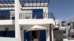 A modern pool area with a clear blue pool, a covered seating area with a table and chairs, and a balcony with blue shutters.