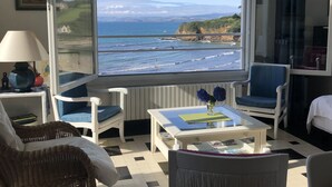A room with a large window overlooking a coastal view, featuring a white table with a vase of flowers, two blue chairs, and a wicker chair.