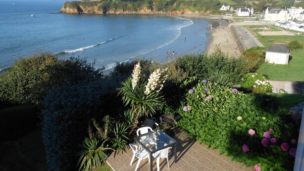 A coastal view with a beach, a small garden with white chairs, and a few houses in the background.