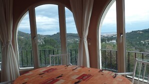 Quarto com mesa redonda, cadeiras brancas e vista para uma paisagem montanhosa através de amplas janelas com cortinas.