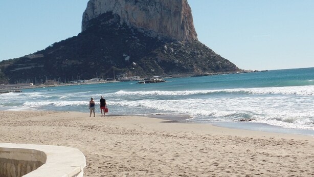 Uma praia com uma rocha imponente ao fundo e duas pessoas caminhando na areia.