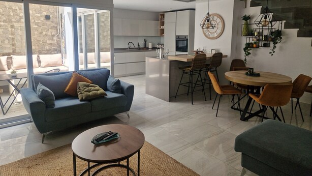 A modern living space with a sofa, coffee table, dining area, kitchen, and large windows.
