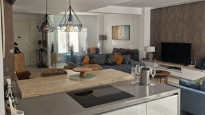 A modern kitchen with a central island, overhead ventilation, pendant lighting, and a living area with a sofa and television.