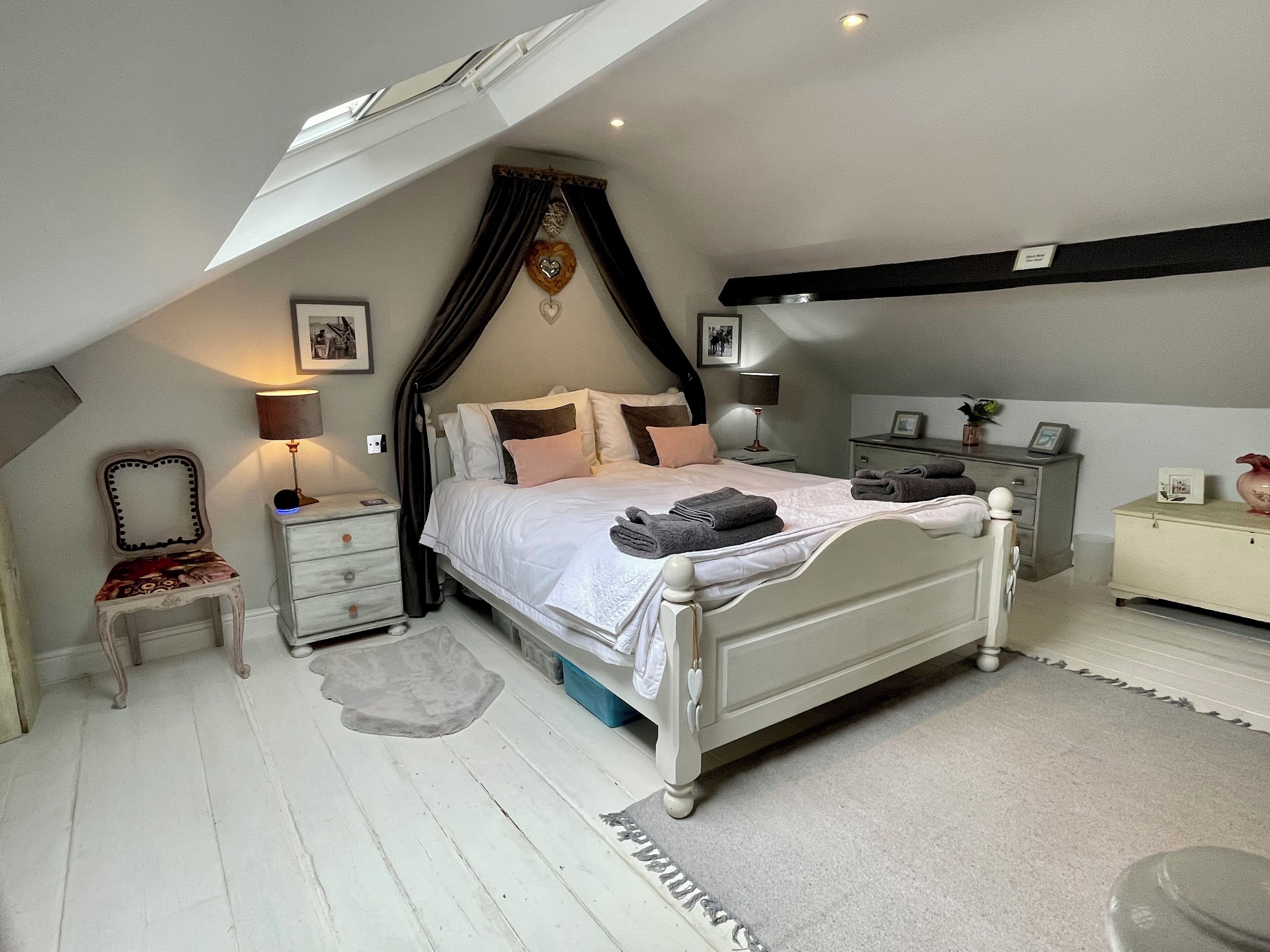 Une chambre avec un plafond en pente, un lit avec une literie blanche, une table de chevet avec une lampe et une commode avec un miroir.