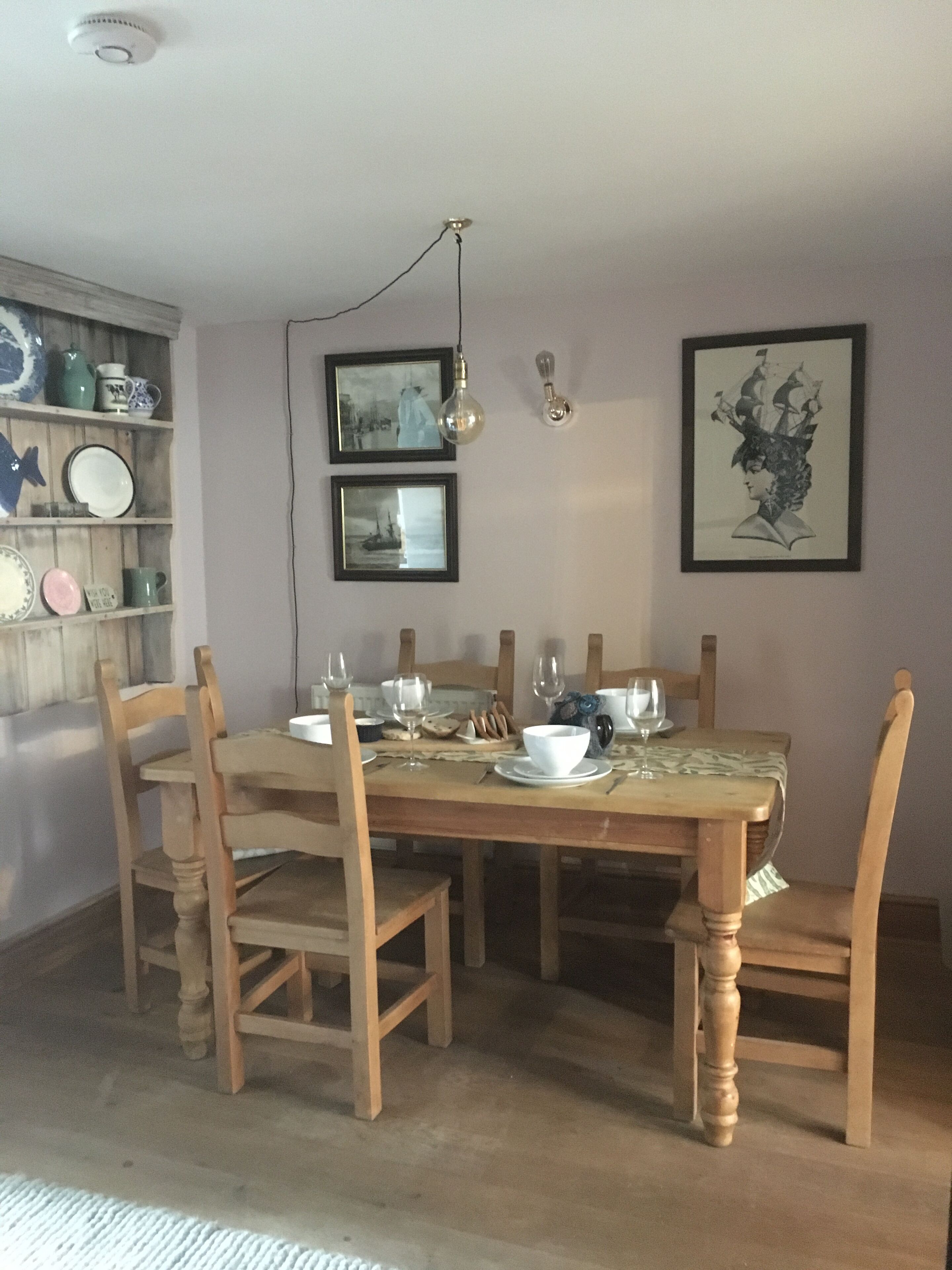 Een eetruimte met een houten tafel voor zes personen, omringd door houten stoelen. Twee ingelijste foto's hangen aan de muur, een plank met verschillende serviesdelen en een hangende lamp.