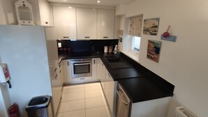 A compact kitchen with white cabinets, a black countertop, and a built-in oven. There is a window with a view of the outside, and a small trash bin is visible. - Pebbles Superb Apartment/Woodland & Sea View/Short Walk Beach/Free Parking &WiFi (Saundersfoot)