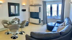 A modern living room with a glass dining table, two chairs, a black leather sofa, a TV, and a window with curtains. - Pebbles Superb Apartment/Woodland & Sea View/Short Walk Beach/Free Parking &WiFi (Saundersfoot)