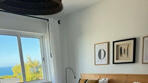A hotel room with two beds, a view of the sea, framed artwork on the wall, and a large window.