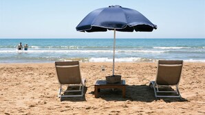 Une plage avec deux chaises longues, une table avec un panier et une bouteille, et un parasol bleu.