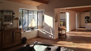 Een ruime woonkamer met een houten plafond, een groot raam met een model zeilboot, een eethoek en een keuken die zichtbaar is door een open doorgang.