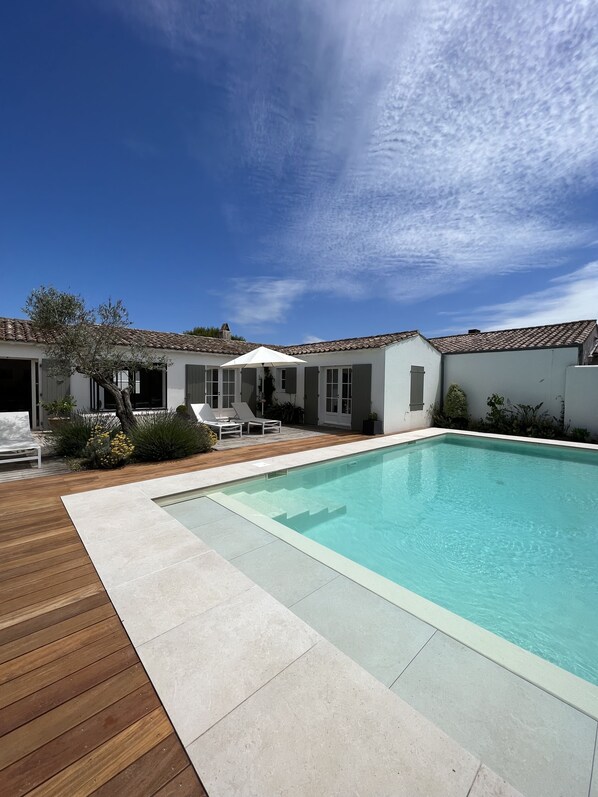 A modern pool area with a wooden deck, a clear blue pool, and a white building with a tiled roof.
