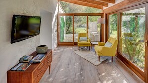 Un saló modern amb un televisor fixat a la paret, un moble de fusta ple de revistes, un butaca groga i una finestra gran amb vista al jardí.