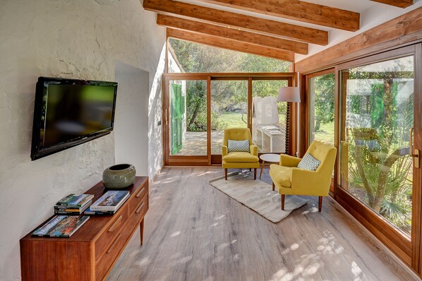 A modern living room with a TV mounted on the wall, a wooden cabinet with magazines, a yellow armchair, and a large window offering a view of the garden.
