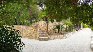 Un camí de grava que condueix a una paret de pedra amb escales, envoltat de diferents plantes i arbres.