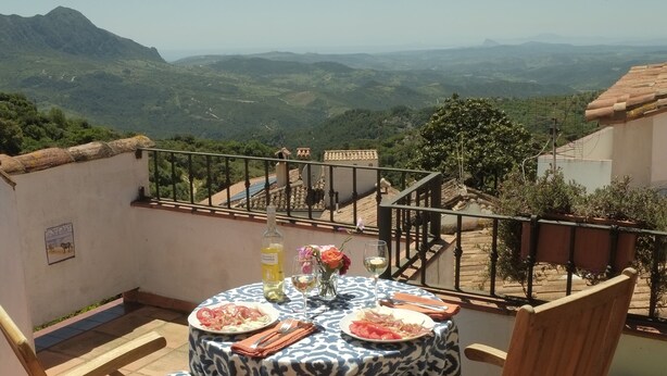 Un tavolo rotondo con tovaglia decorata, due sedie di legno e una bottiglia di vino su una terrazza con vista sulle montagne lontane.