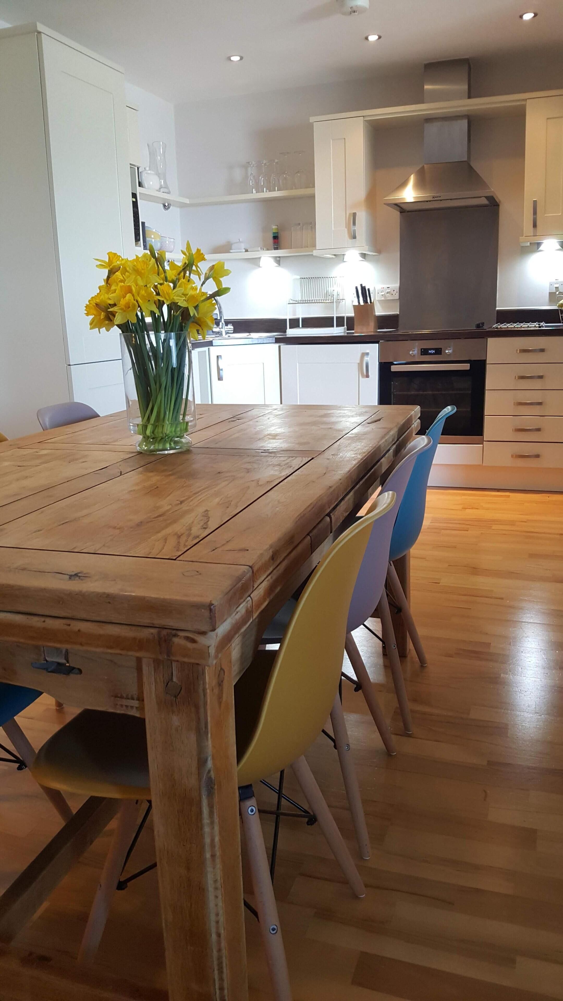 Una mesa de comedor de madera con sillas, un jarrón de flores amarillas y una zona de cocina con armarios y estufa.