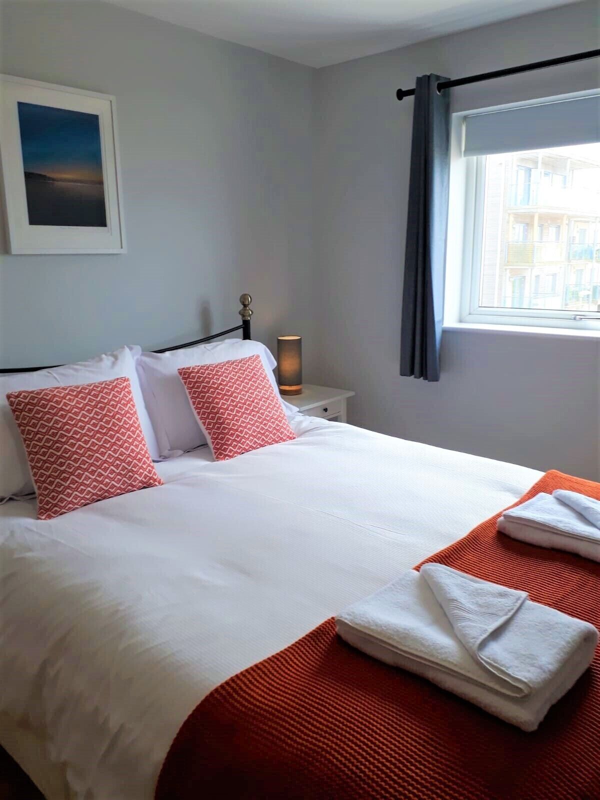 Una cama bien hecha con almohadas blancas y rojas, un plaid rojo y toallas dobladas sobre una mesita de noche blanca con una lámpara.