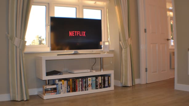 A television on a white stand with a bookshelf below it, a door to the right, and a window with curtains to the left.