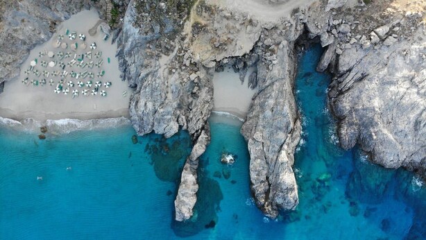 An aerial view of a coastal area with rocky cliffs, clear blue waters, and a sandy beach with numerous umbrellas. - Villa Ostria, Lefkogia Cottage on a 20.000 sq.m.farm! Only 1 km from the beach.  (Rethymno)