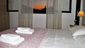 A hotel room with a bed, a window with a view of the sunset, and a nightstand with a lamp. - Villa Ostria, Lefkogia Cottage on a 20.000 sq.m.farm! Only 1 km from the beach.  (Rethymno)