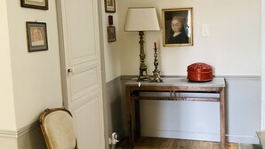 Una habitación con piso de madera, una silla, una mesa con lámpara y cuadros en la pared.