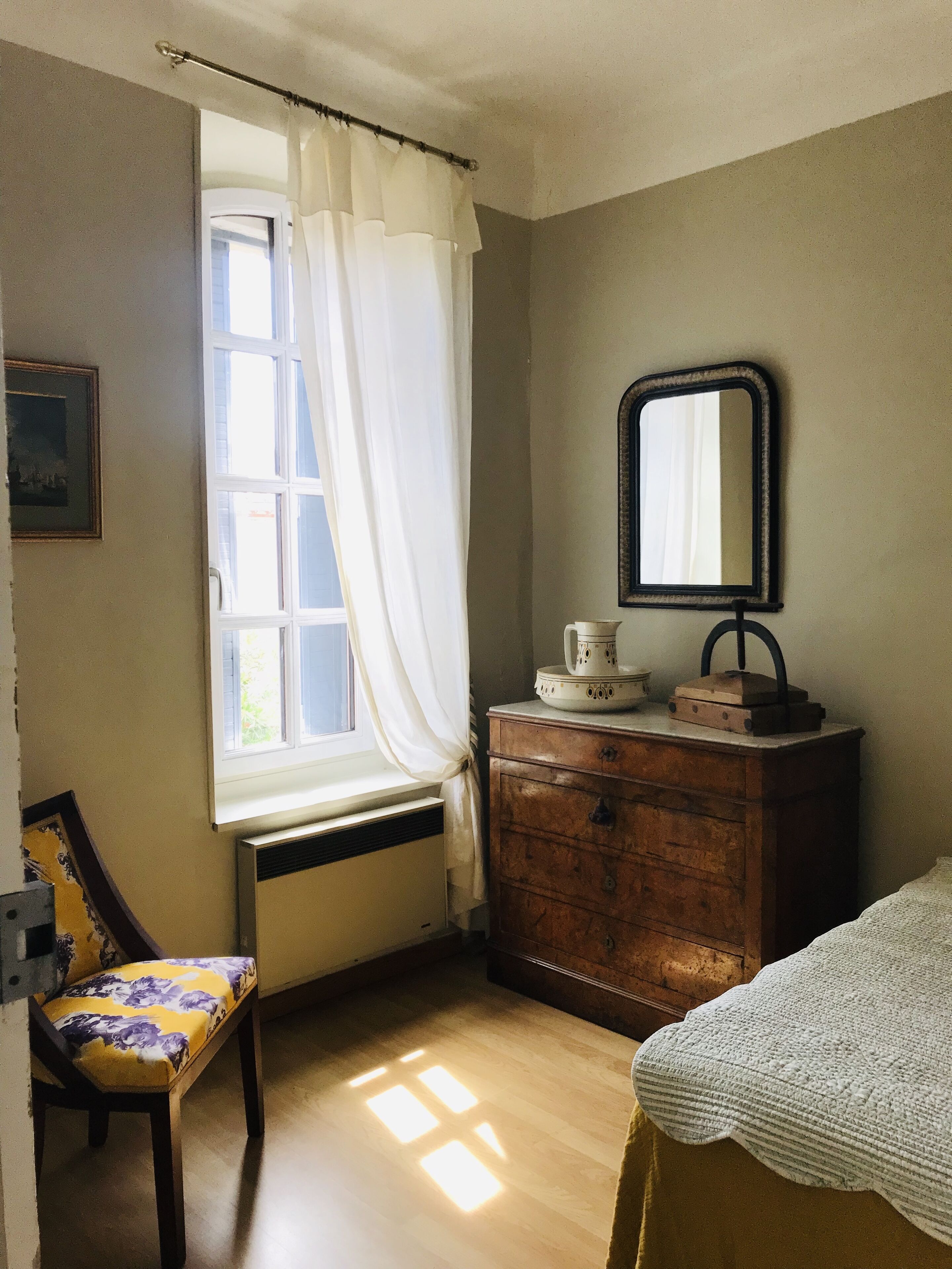 Una habitación con ventana, tocador de madera, una silla y una cama.