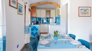 Un comedor con mantel azul, platos blancos y sillas azules. Se ve una cocina al fondo con azulejos azules y armarios blancos.